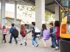 Elementary school students wearing backpacks line up to enter the building next to a yellow school bus.