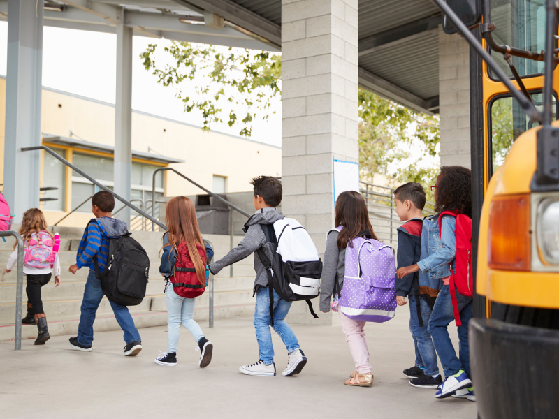 Elementary school students wearing backpacks line up to enter the building next to a yellow school bus.