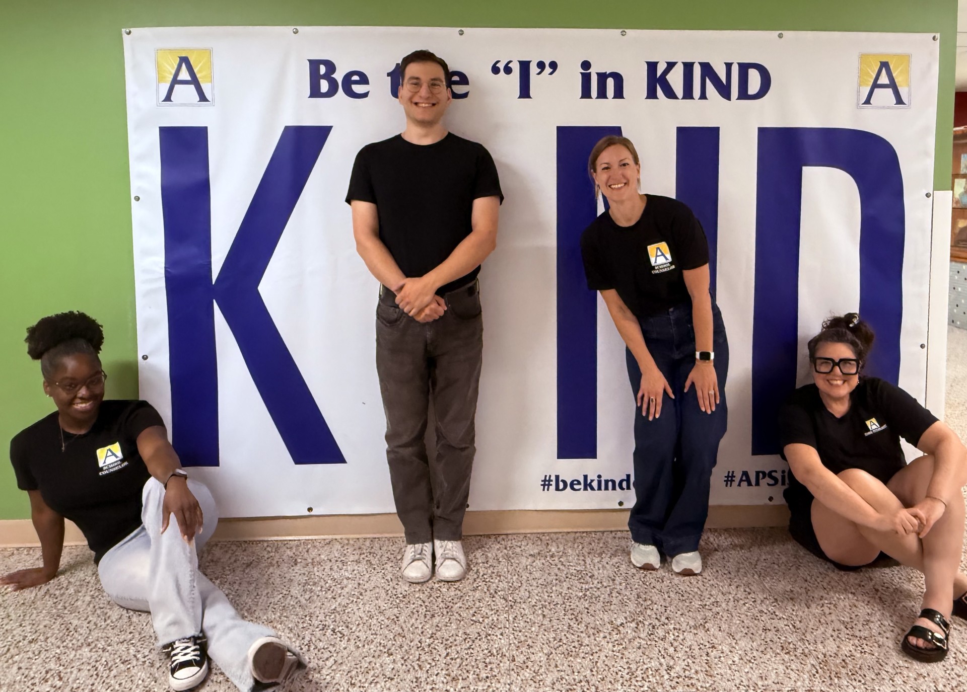 Four school staff members pose in front of a large “Be the ‘I’ in KIND” banner, wearing matching black Ashlawn Elementary shirts and smiling at the camera.