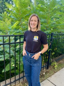 A smiling school counselor stands outdoors in front of lush green foliage and a black metal fence, wearing a black school counselor T-shirt and blue jeans.